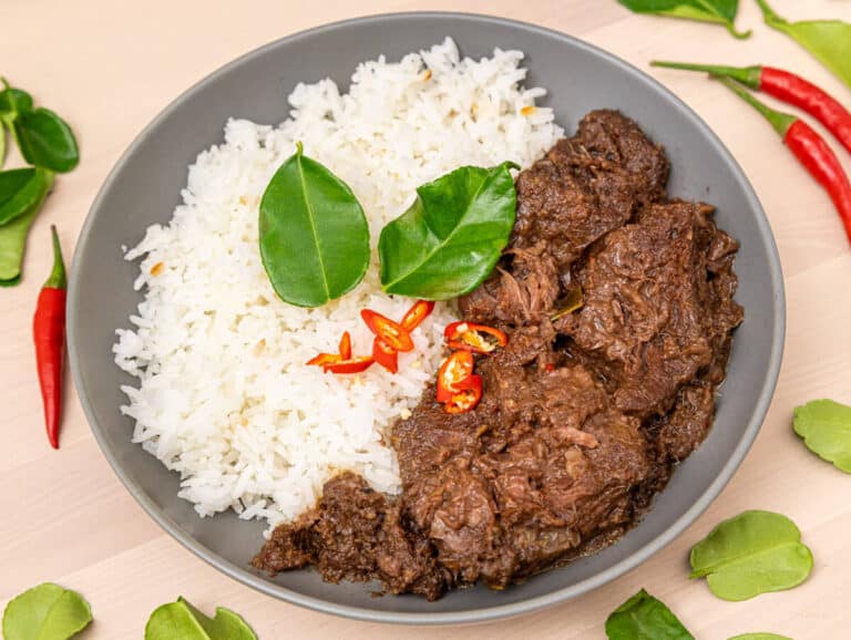 Plate of fragrant rice with tender beef rendang, garnished with red chilies and fresh lime leaves, on a wooden table.